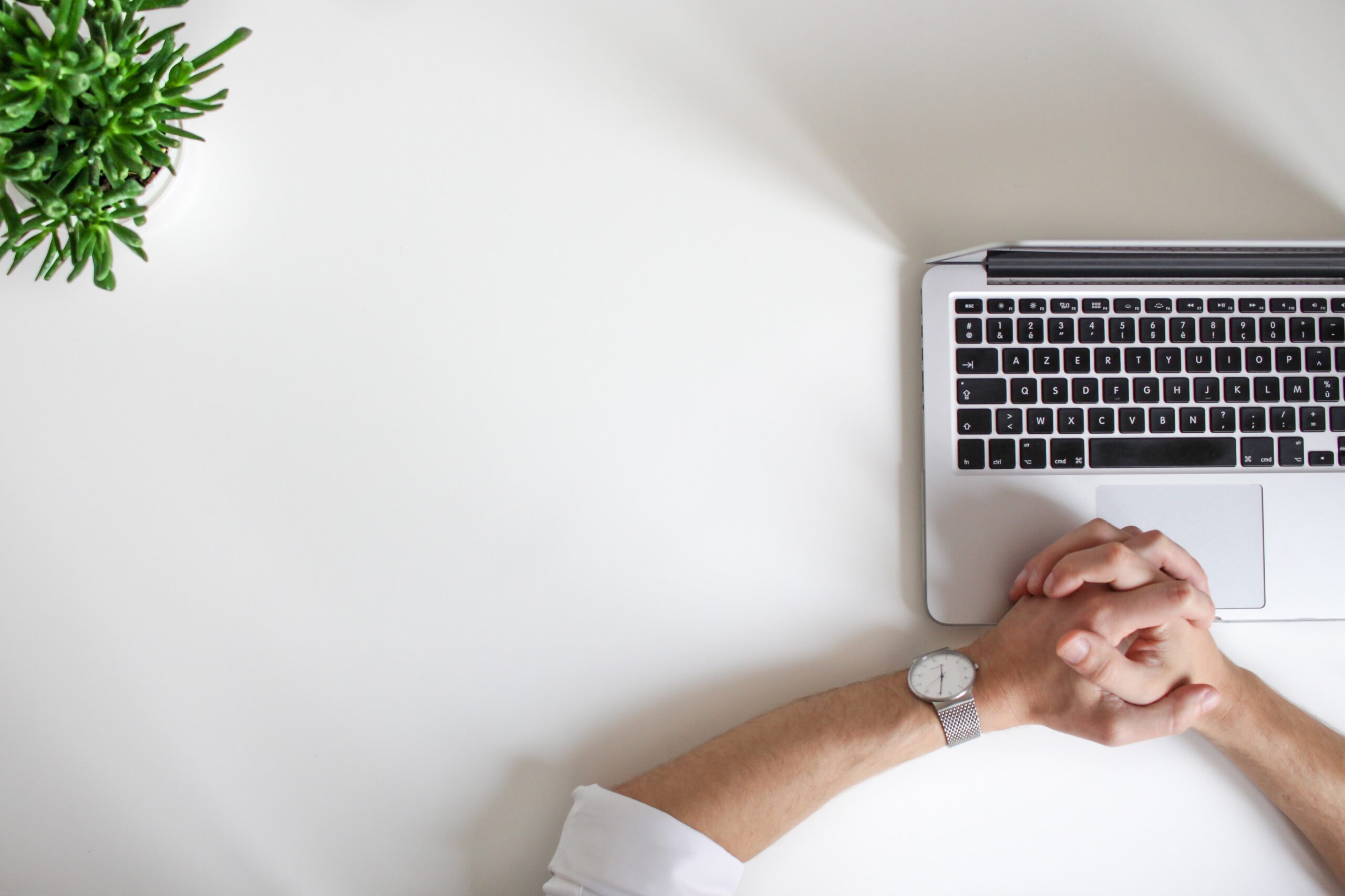 Two hands clasped together on a laptop, and the laptop is sitting on a desk with a plant in the corner of the photo.