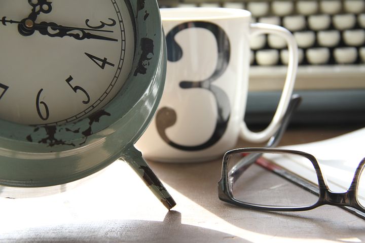 A photo of a teal clock, a mug with the number 3 on it, a pair of glasses, and a typewriter in the background.